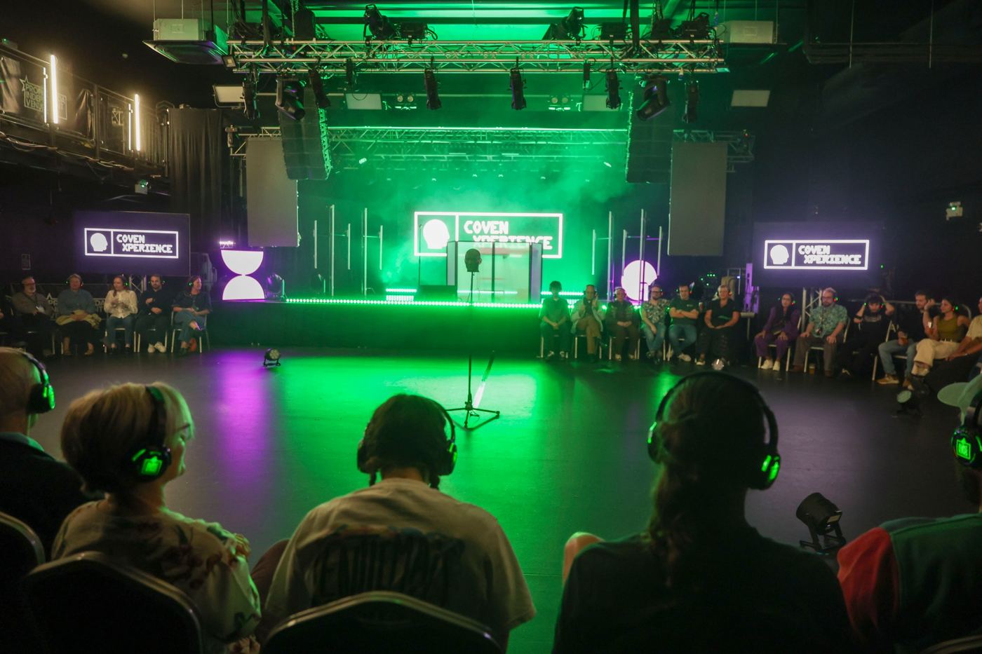 Immersive audio experience: audience in silent-disco headsets seated in the round under green stage lighting